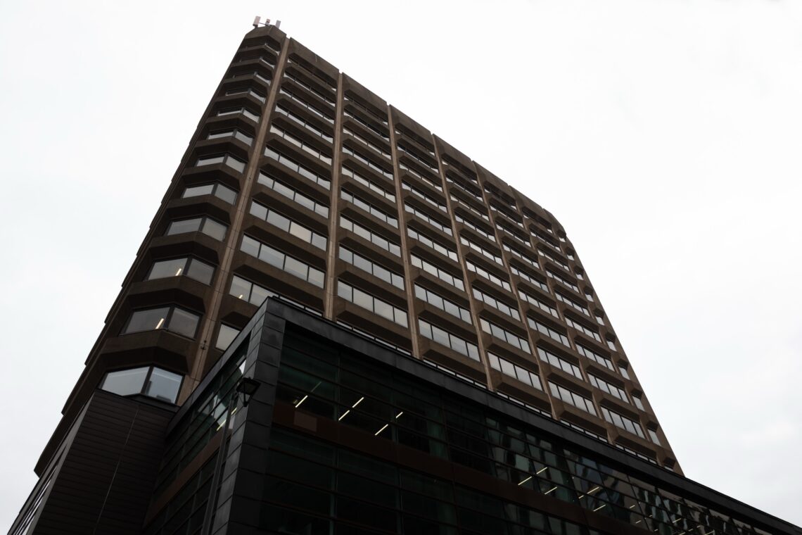 A tall, modern office building with dark-tinted windows and geometric lines rises against an overcast sky, viewed from a low angle. The structure features a mix of concrete and glass on its angular façade.