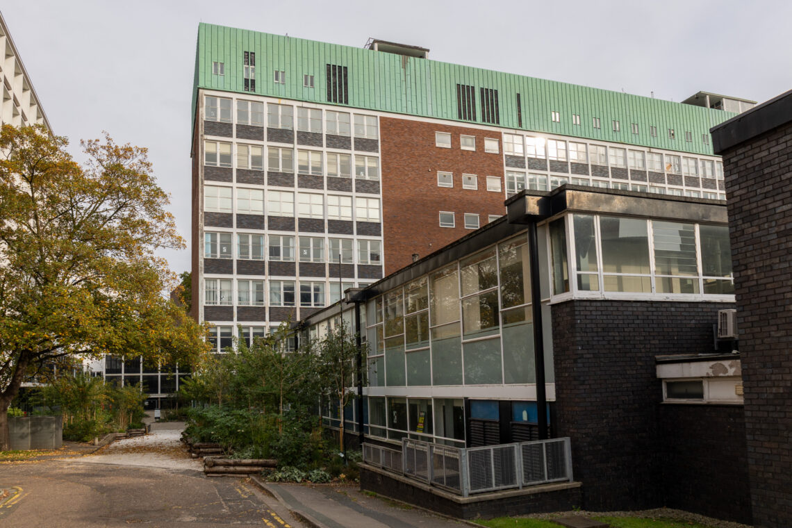 A multi-storey building with a green roof, red brick and glass facade stands behind a smaller, dark brick building. Trees and shrubs grow nearby along a paved path under a grey sky.