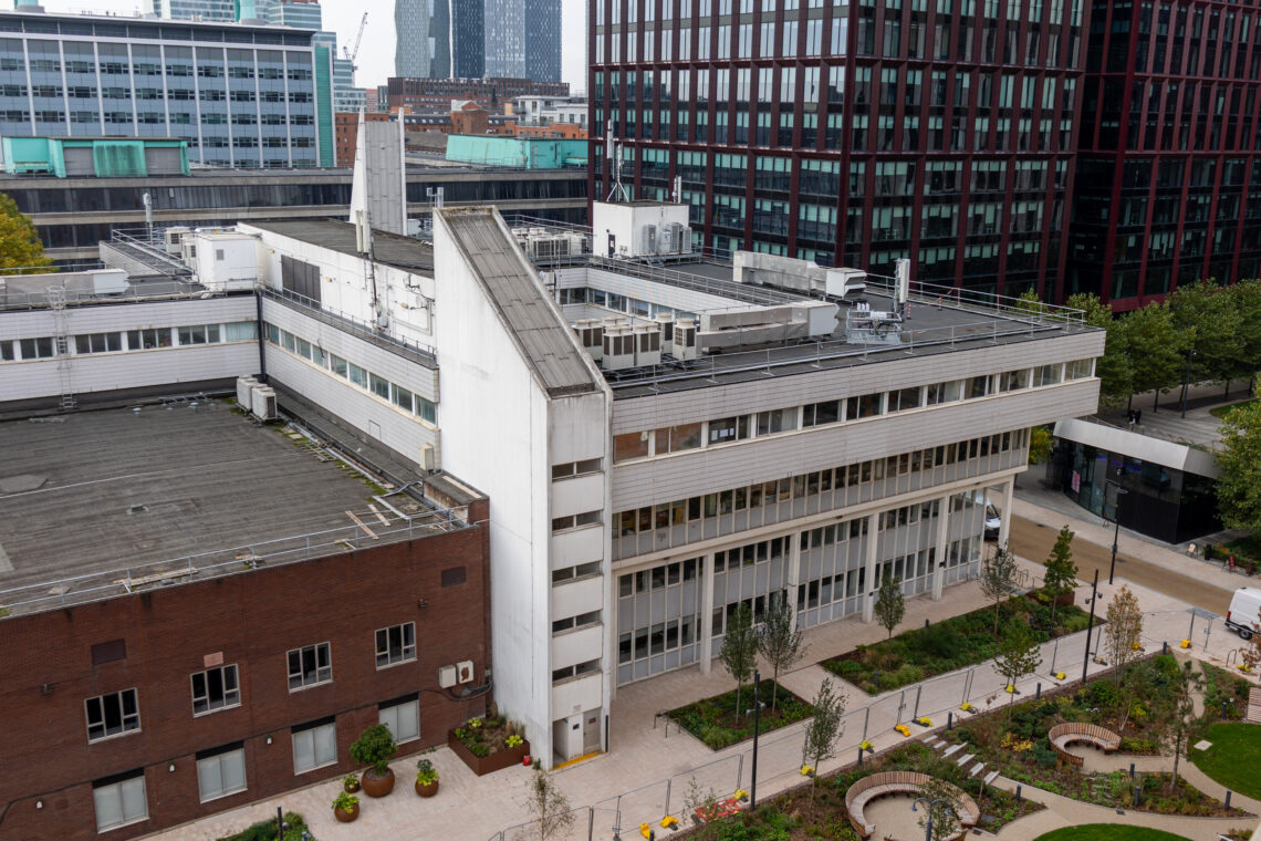 Aerial view of a modern white building with large windows, rooftop equipment, and adjacent brown structure. The foreground features a landscaped garden with trees, bushes, curved pathways, and benches. Tall glass office buildings rise in the background.