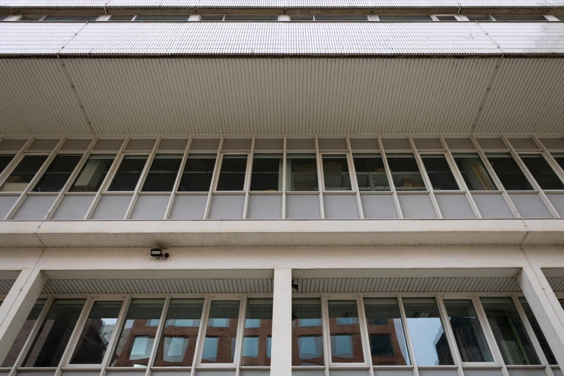 Low-angle view of a modern, white façade building with large rectangular windows and vertical columns. Reflections of nearby buildings are visible in the glass. The structure has a geometric and minimalist architectural style.