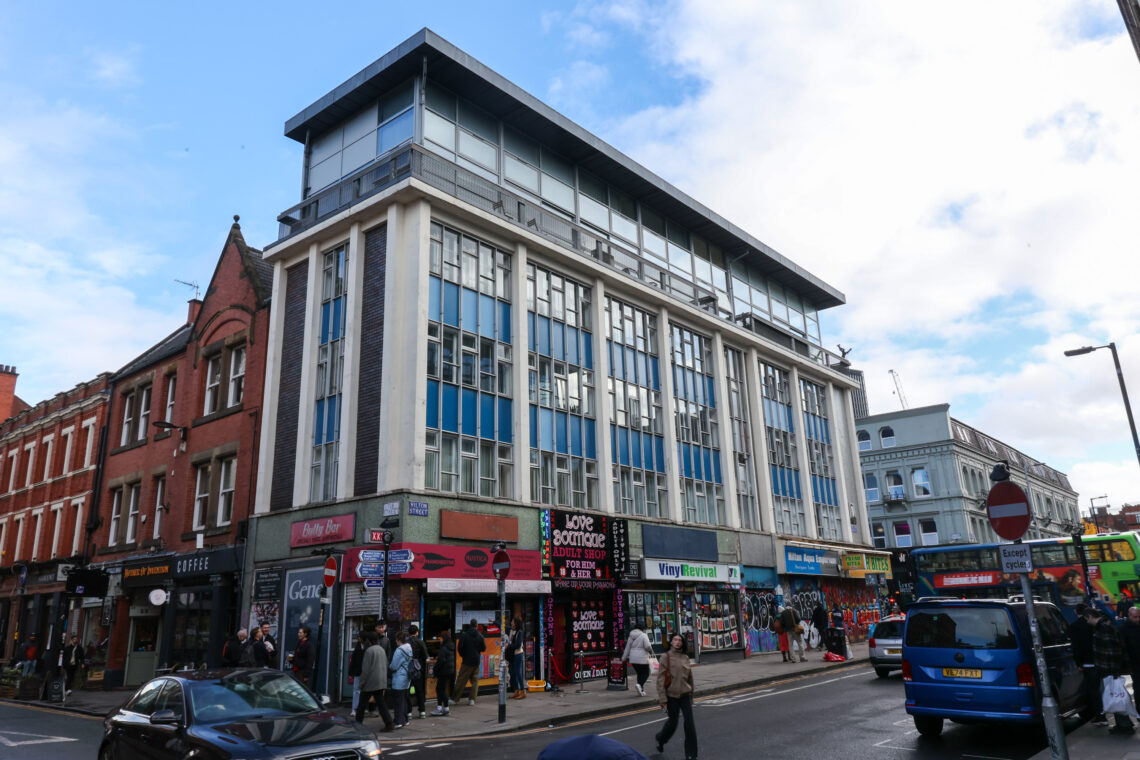 A busy street scene with people walking past shops, including a comic shop and a record shop, at the base of a modern building with large blue and white windows. Older brick buildings and parked cars line the street under a partly cloudy sky.