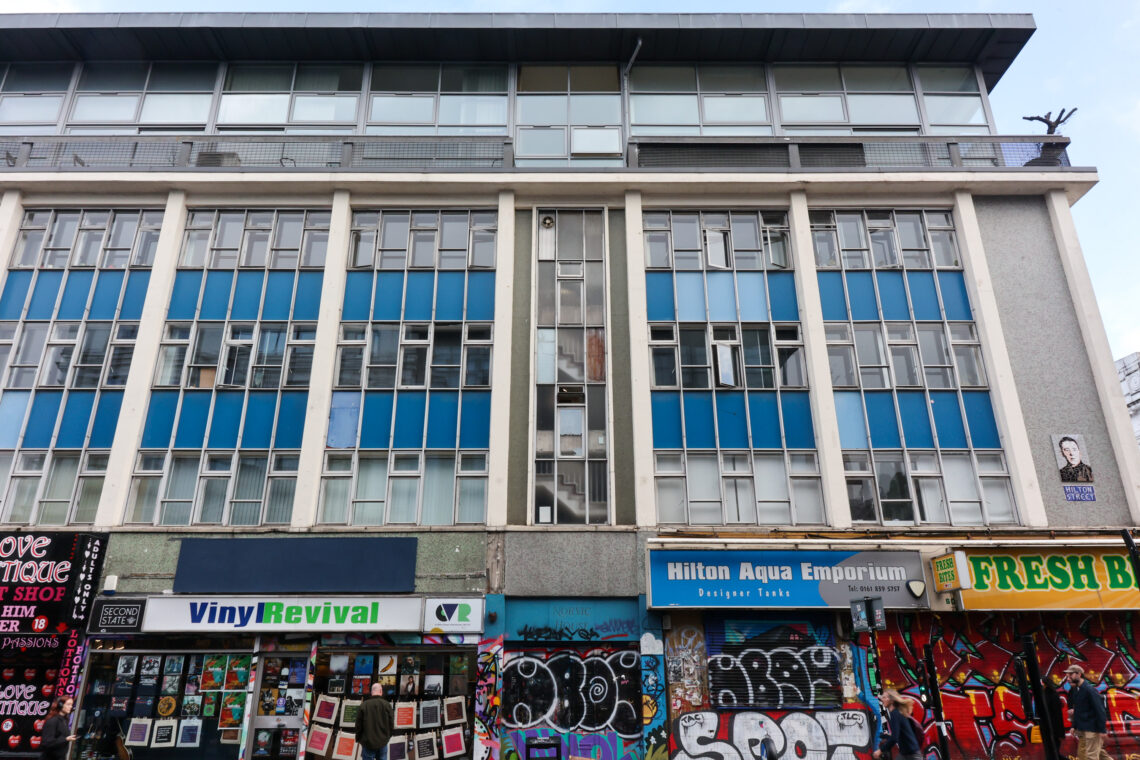 A three-storey building with blue and white panels above colourful graffiti-covered shopfronts, including Vinyl Revival and Hilton Aqua Emporium. The upper floors have many windows, and a small statue stands on the roof’s far right corner.