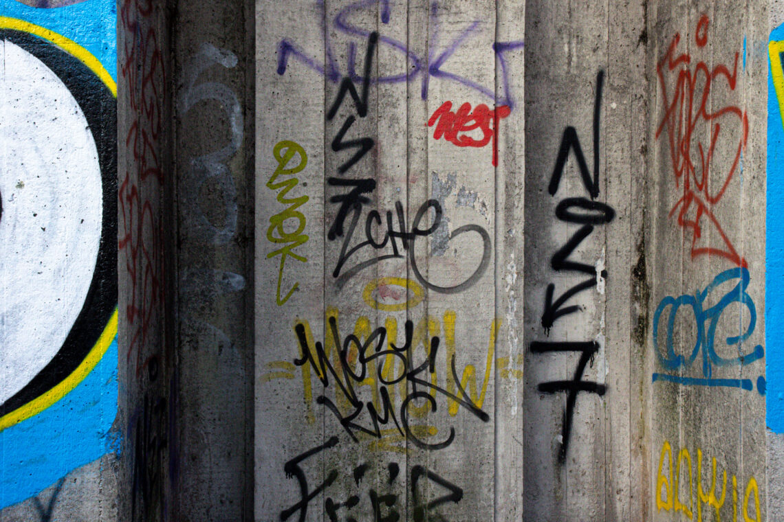 A concrete pillar covered in various colourful graffiti tags and spray-painted words, mostly in black, yellow, and red. On the left, a mural features a partial white circle outlined in blue and yellow, contrasting the rough, layered tags on the pillar.