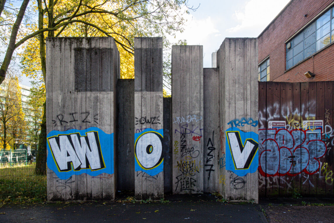 Four tall concrete slabs with blue-bordered white graffiti letters “AW,” “O,” and “V.” Additional colourful tags and graffiti cover the slabs. Trees and a fence are on the left; a brick building with windows is on the right. Sunlight filters through leaves.