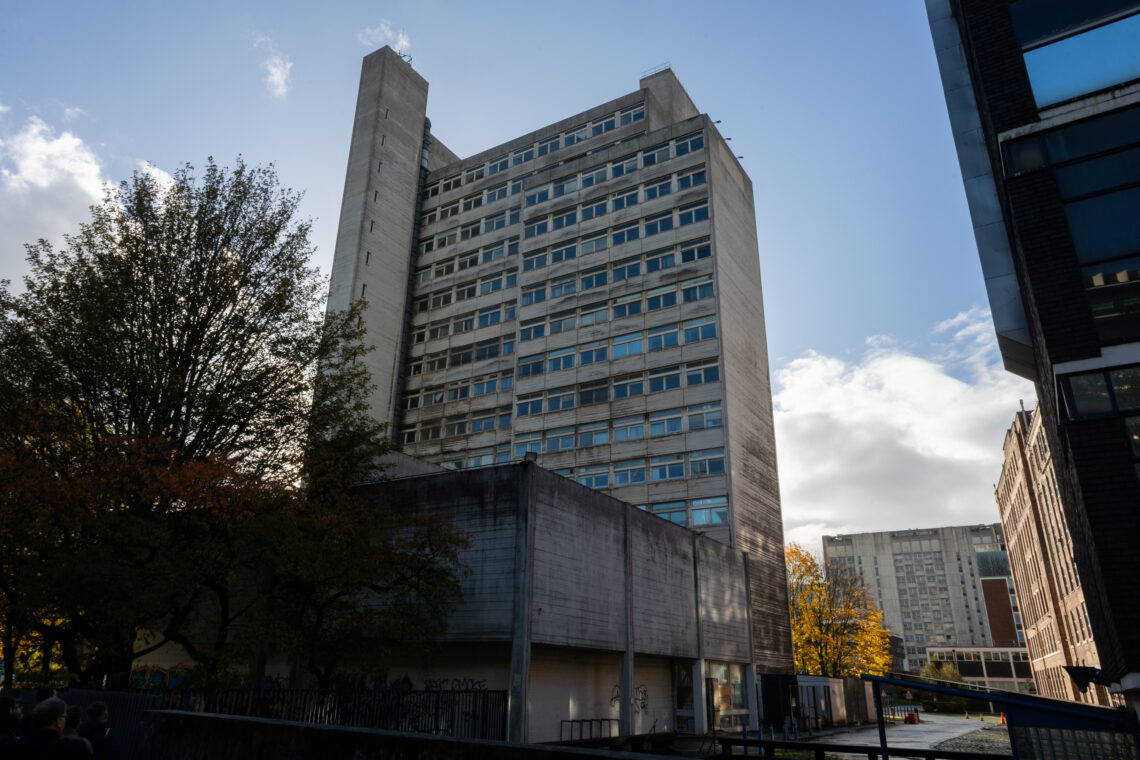 A tall, grey concrete block of flats with many windows stands between trees and other buildings. The sun is shining behind clouds, casting shadows. Autumn leaves are visible on the trees, and the sky is mostly blue with some clouds.
