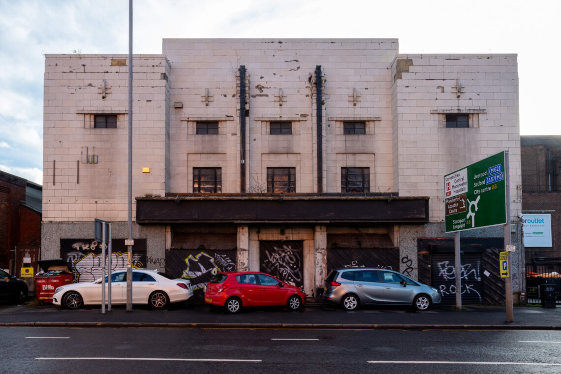 A derelict, white Art Deco building with boarded-up windows and graffiti on its facade. Four cars are parked in front. A street sign and lamp post are visible on the pavement. The sky is partly cloudy.