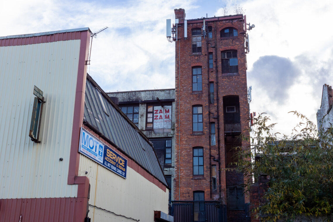 A tall, old brick building with a faded ZAM ZAM sign stands behind a white MOT service garage. The sky is cloudy, and green tree branches are visible on the right side. The buildings show signs of age and wear.