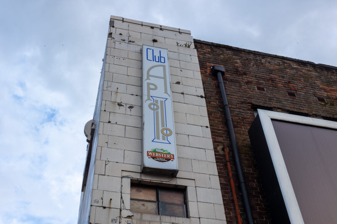A tall, white tiled building facade features a vintage vertical sign reading Club Apollo. Below the sign is a small Werstecks beer advert. The building next to it has red-brown bricks. The sky above is cloudy.