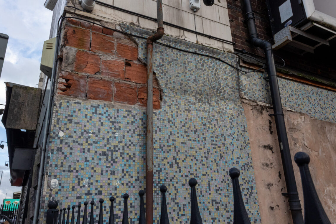 A weathered building wall shows damaged red bricks, cracked plaster, and faded blue mosaic tiles. Pipes run vertically, and a black metal fence is in the foreground. Electrical boxes and vents are visible near the top. Sky and buildings appear in the background.
