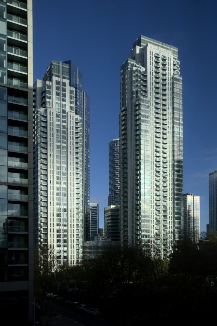 Tall modern glass skyscrapers dominate the scene under a clear blue sky, reflecting sunlight. Trees and a few low-rise buildings are visible at the bottom, with shadows cast on the street below. Urban cityscape creates a striking contrast with the sunlight.