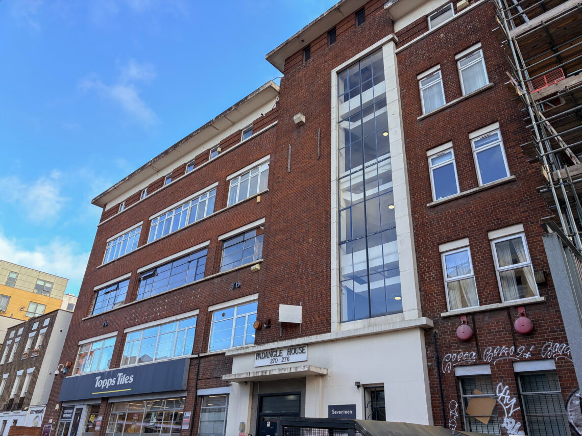 A red-brick office building has many large windows, a glass stairwell, and signs for Topps Tiles. There’s a construction scaffold on the right. The sky is clear and blue.