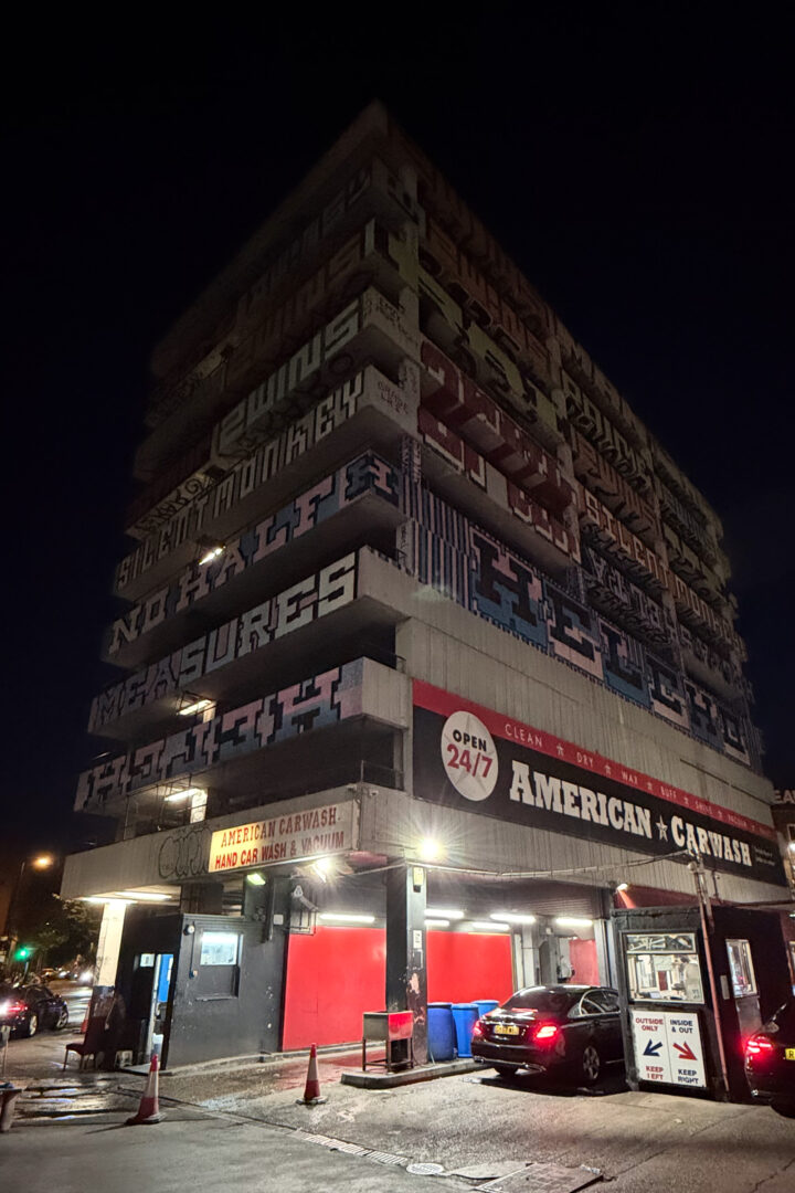 A multi-storey building at night features bold graffiti-style text on each balcony. The ground floor houses an American car wash with red, black, and white signage, brightly lit. Several cars are parked nearby under streetlights; the sky is dark.