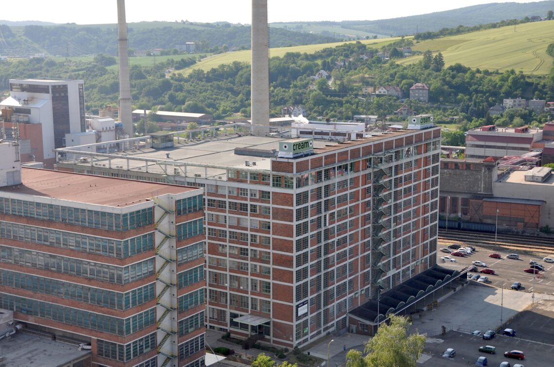 A large industrial brick building with many windows stands near two tall chimneys. Car parks and cars surround it. Green hills, trees, and scattered houses fill the background under a clear sky.