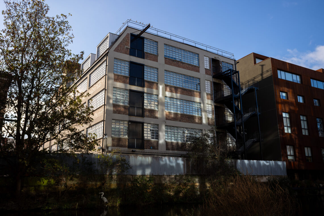 A large, white and brown industrial building with many grid-like windows stands beside a modern brown building. Trees and bushes are in the foreground, and metal stairs run up the building’s side. The sky is clear and blue.