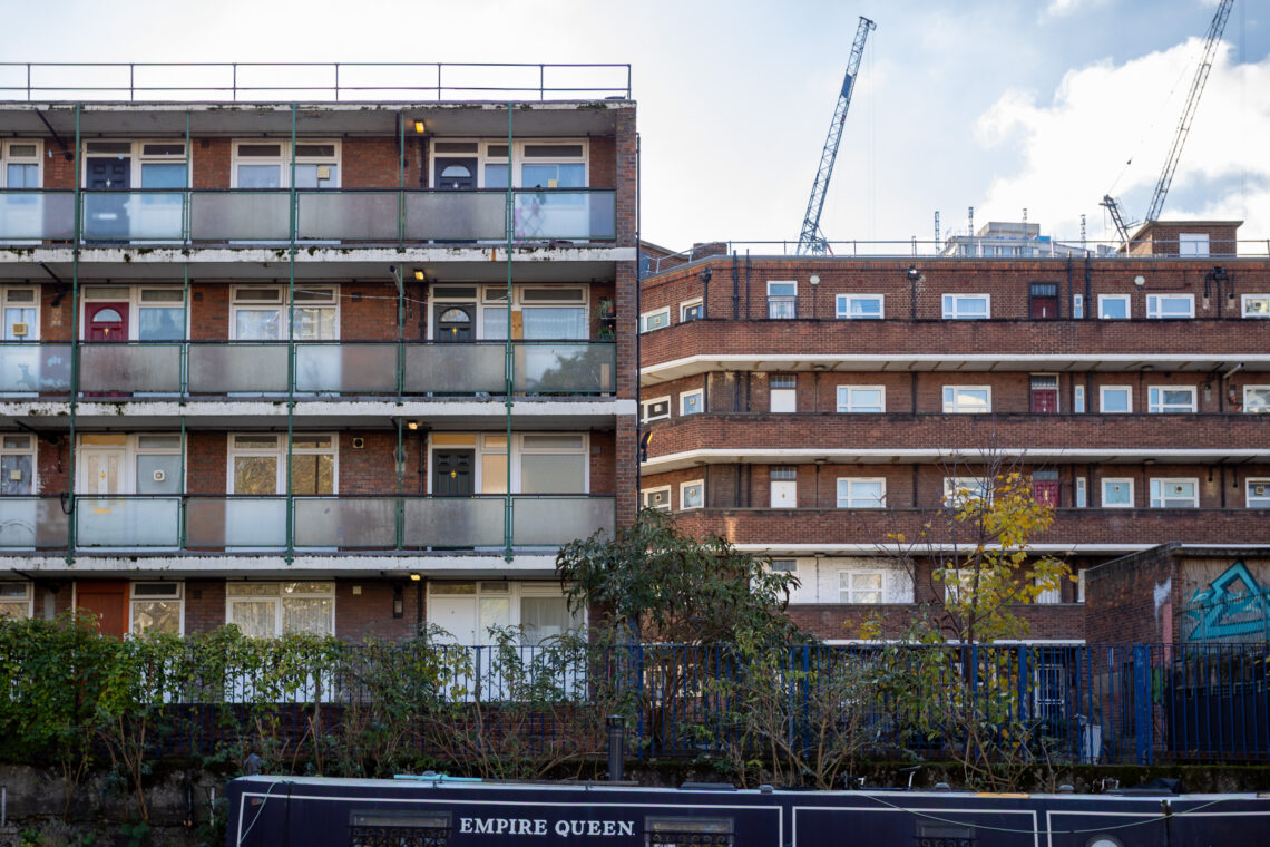 A canal boat labelled EMPIRE QUEEN is in front of two brick blocks of flats with balconies. Cranes are visible in the background, and plants grow along the fence between the boat and the buildings. The sky is partly cloudy.