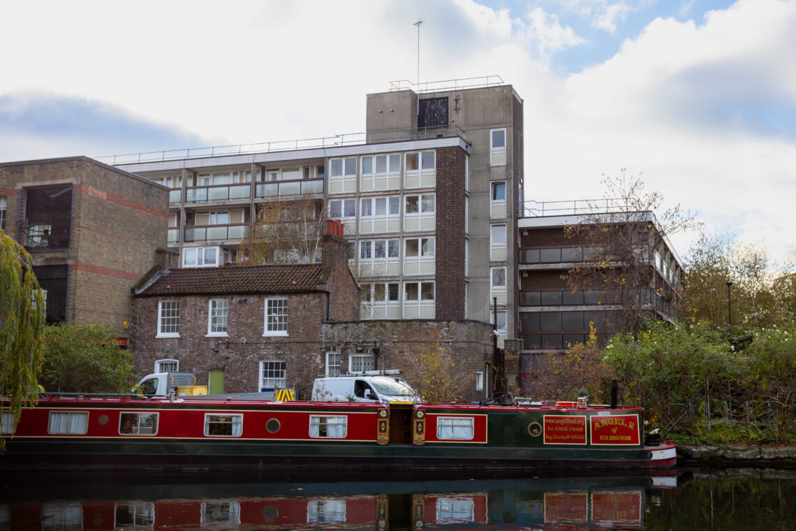 A red canal boat floats on a calm canal in front of a two-storey brick cottage and a tall, modern block of flats. Trees with autumn leaves border the water, and a white van is parked beside the cottage. The sky is cloudy.