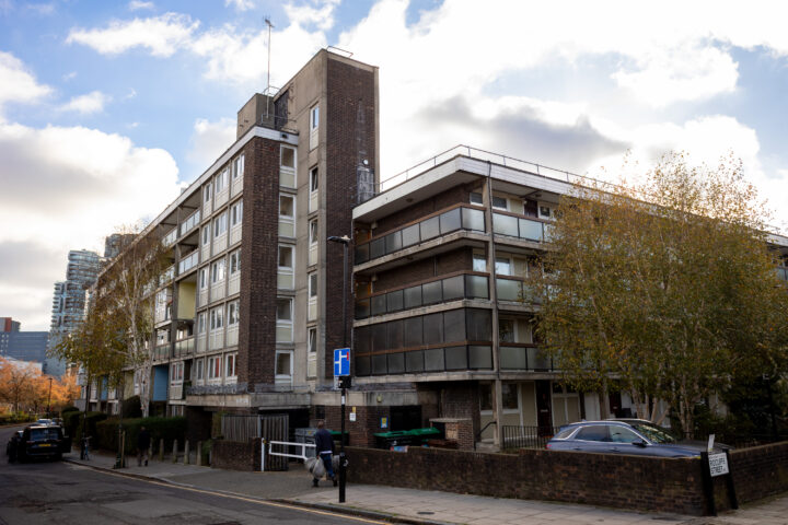 A brown brick block of flats with balconies and a blue hospital sign is seen on a sunny day. Parked cars line the street, and trees with autumn leaves stand nearby. The sky is partly cloudy, and a tall, modern building is visible in the background.
