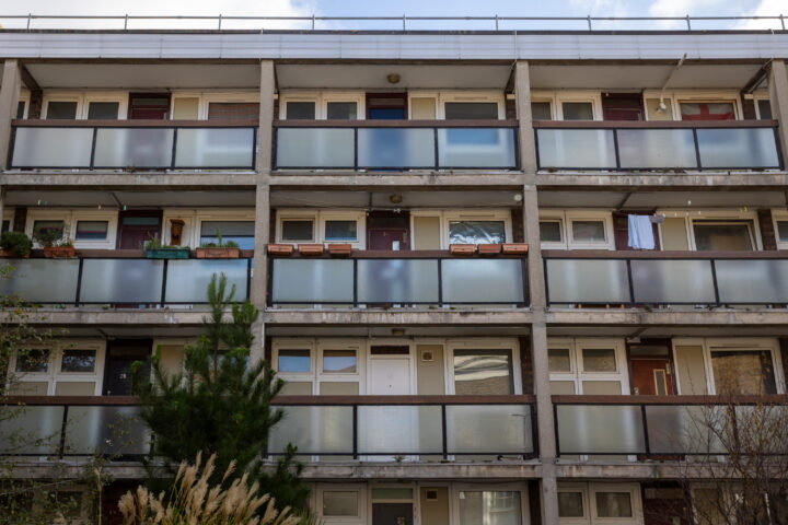A three-storey block of flats with frosted glass balcony railings, white doors, and windows. Some balconies have planters, potted plants, or washing hanging. There is greenery, including a pine tree, in front of the building. The sky is partly cloudy.