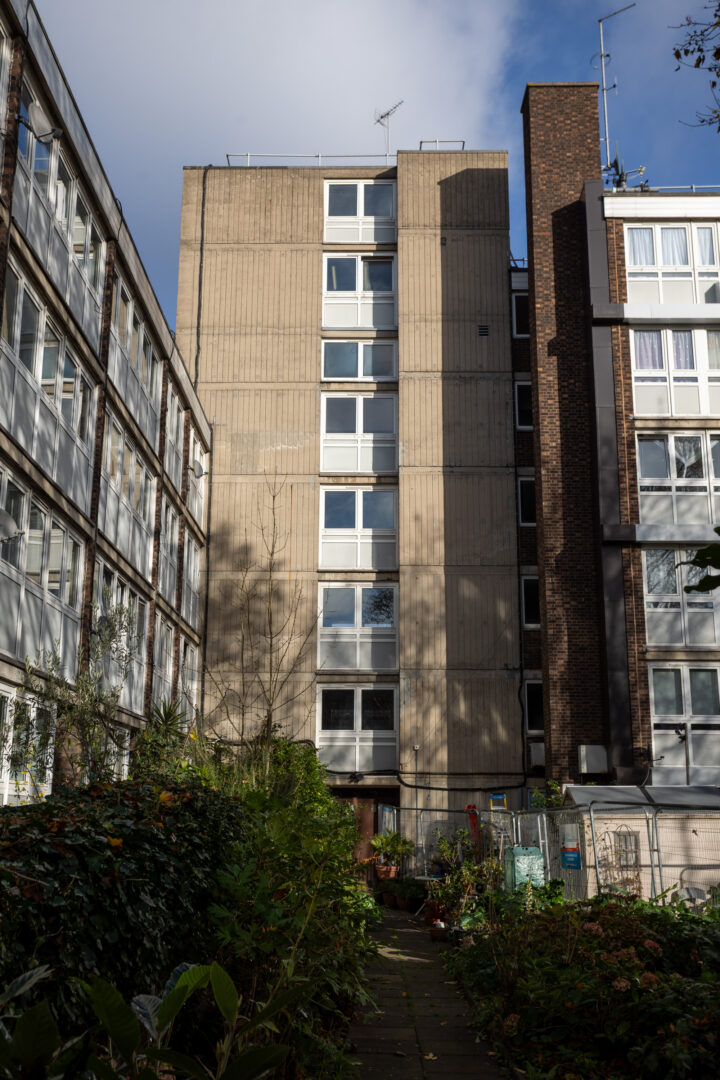 A tall, plain concrete block of flats with white-framed windows stands between two other buildings. The entrance is at the bottom, surrounded by greenery, overgrown plants, and a small fenced-off area in the foreground. The sky above is partly cloudy.