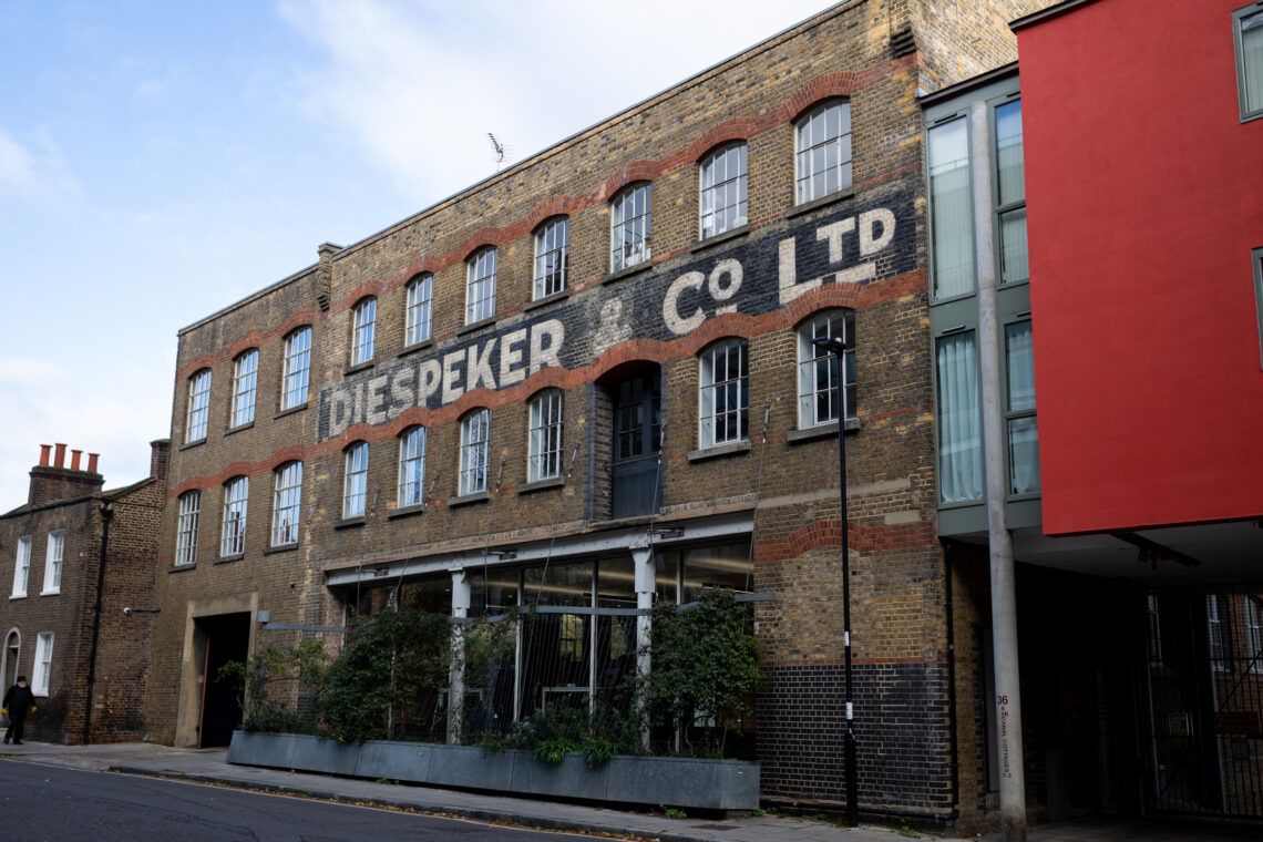 A three-storey brick building with DIESPEKER CO LTD painted in large white letters, featuring arched windows, a main entrance with glass doors, and greenery in front. The sky is partly cloudy, and a modern building adjoins it on the right.