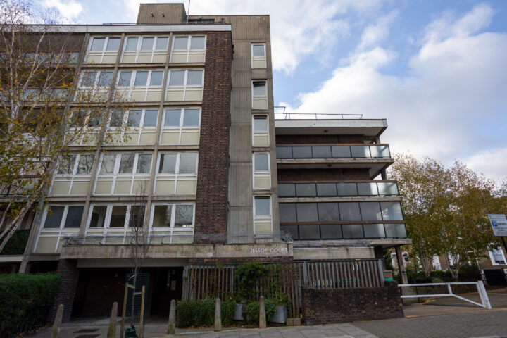 A five-storey block of flats with beige and brown brick sections, large windows, and balconies. Leafy trees stand nearby. The sign reads Assup Court. The sky is partly cloudy and there’s a paved pavement in front.