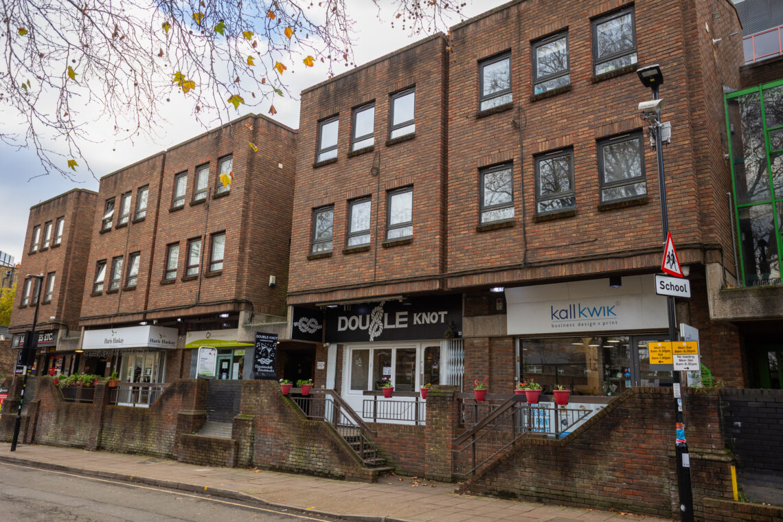 Three-storey brick building with shops at street level, including a hair salon, Double Knot, and Kall kwik. Stairs lead to entrances. Leafless tree branches overhang above. A School road sign is visible. Windows reflect trees. The pavement is mostly empty.