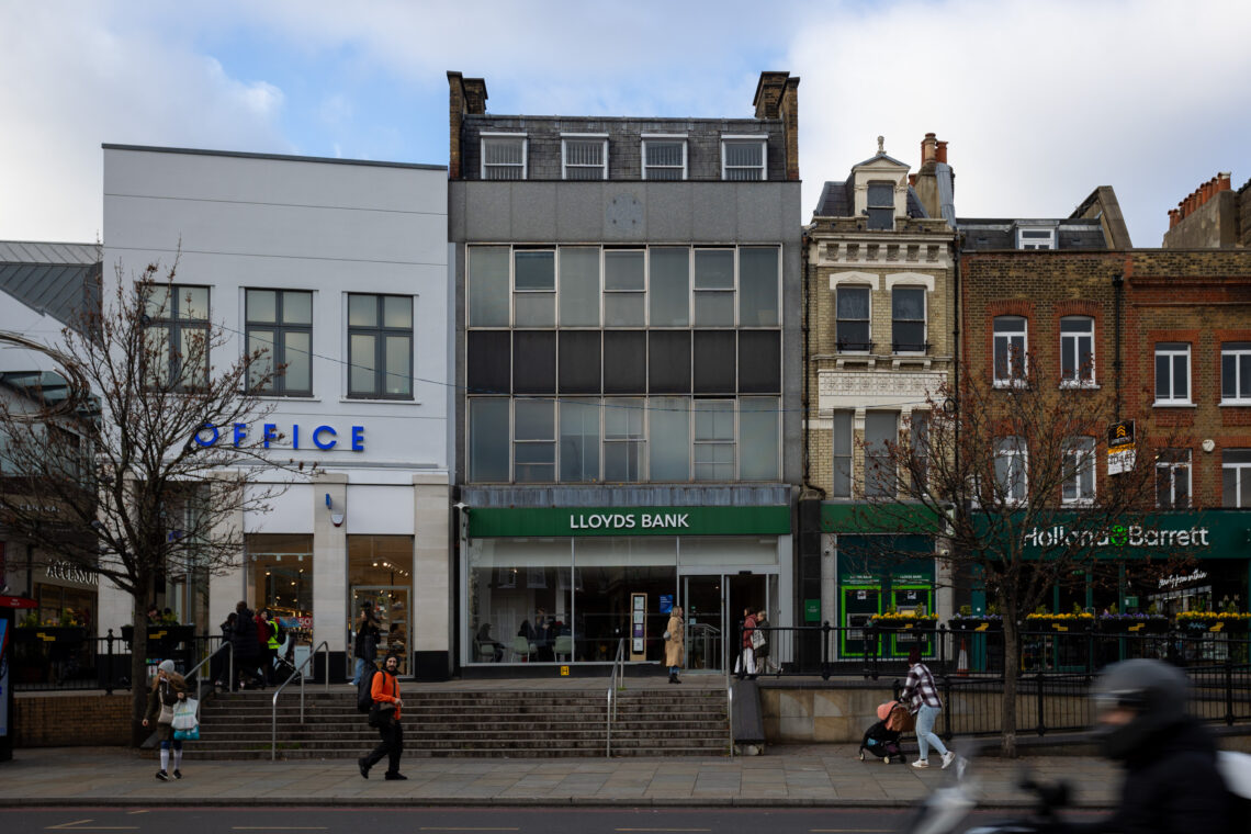 Three city buildings: a white OFFICE shoe shop, a grey LLOYDS BANK with glass doors and steps, and a brown-brick Holland & Barrett. People walk by on the pavement; leafless trees stand in front. Sky is cloudy.