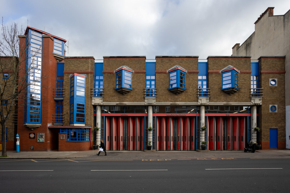 Modern brick fire station with five red-and-white garage doors, blue window frames, projecting blue-finished windows above, and vertical blue glass panels on the left. Two people walk outside on the pavement; a bare tree stands to the left of the entrance.