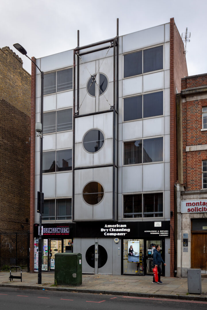 A four-storey modern building with circular windows, metallic panels, and a central frame. Ground level has shops including American Dry Cleaning Company. Two people stand outside; a street lamp and kiosk are visible.