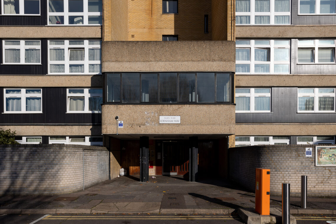 A modern block of flats entrance with a concrete overhang, large windows above, brick and grey panel facade, and a sign reading Edenham Way. An orange parking barrier is visible in the foreground.