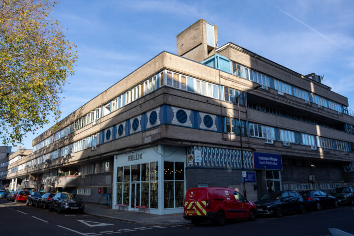 A mid-century modern building with geometric patterns, large windows, and shops, including RELLIK, on a sunny street. Cars are parked along the kerb, and a red van is in the foreground. A leafy tree stands at the left, and a blue sky is above.