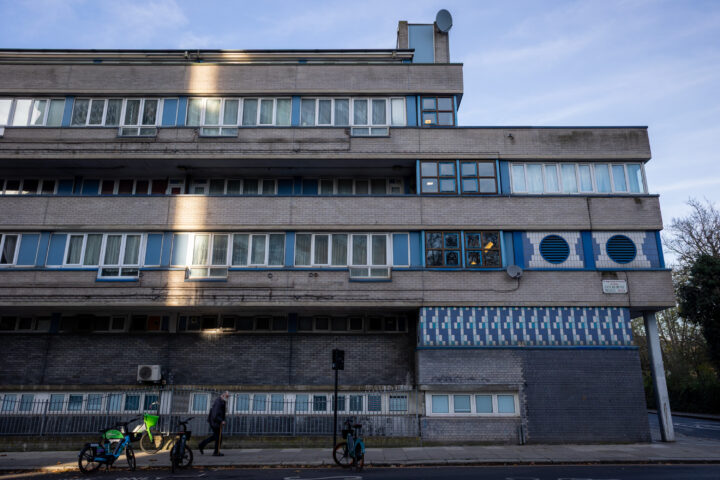 A person walks past a modern, grey brick block of flats with horizontal windows and blue accents. Bicycles are parked along the pavement. A shaft of sunlight highlights part of the building. The sky is clear and blue.