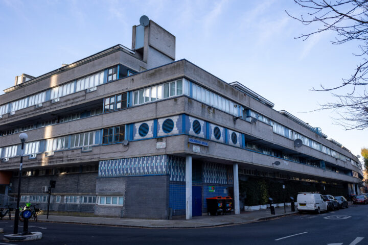 A three-storey, modernist building with long horizontal windows, blue tiles, and geometric designs stands at a street corner. Several cars are parked beside it. A person in a high-visibility vest is near a bicycle rack. Trees frame the image on the right.