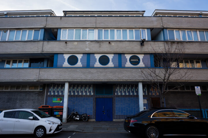 A grey and blue modern block of flats with horizontal windows, round vents, and blue tile detailing. Parked cars and a motorbike line the street. A leafless tree stands near the pavement under a bright, clear sky.