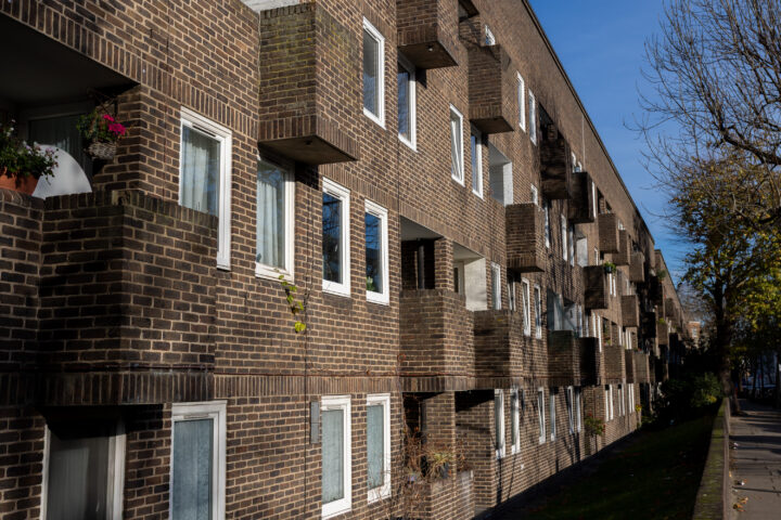 A long, three-storey brick block of flats with small balconies and white-framed windows faces a tree-lined pavement on a sunny day. Some balconies have plants, and shadows suggest it is morning or late afternoon.