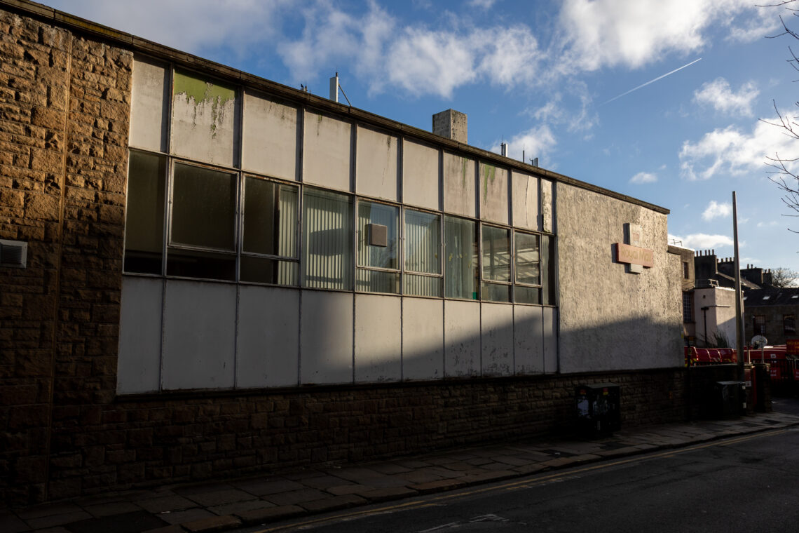 A sunlit, old building with large, grimy windows, white panels, and a stone base lines a quiet street with bins and shadows. The sky is blue with clouds and a visible plane vapour trail.