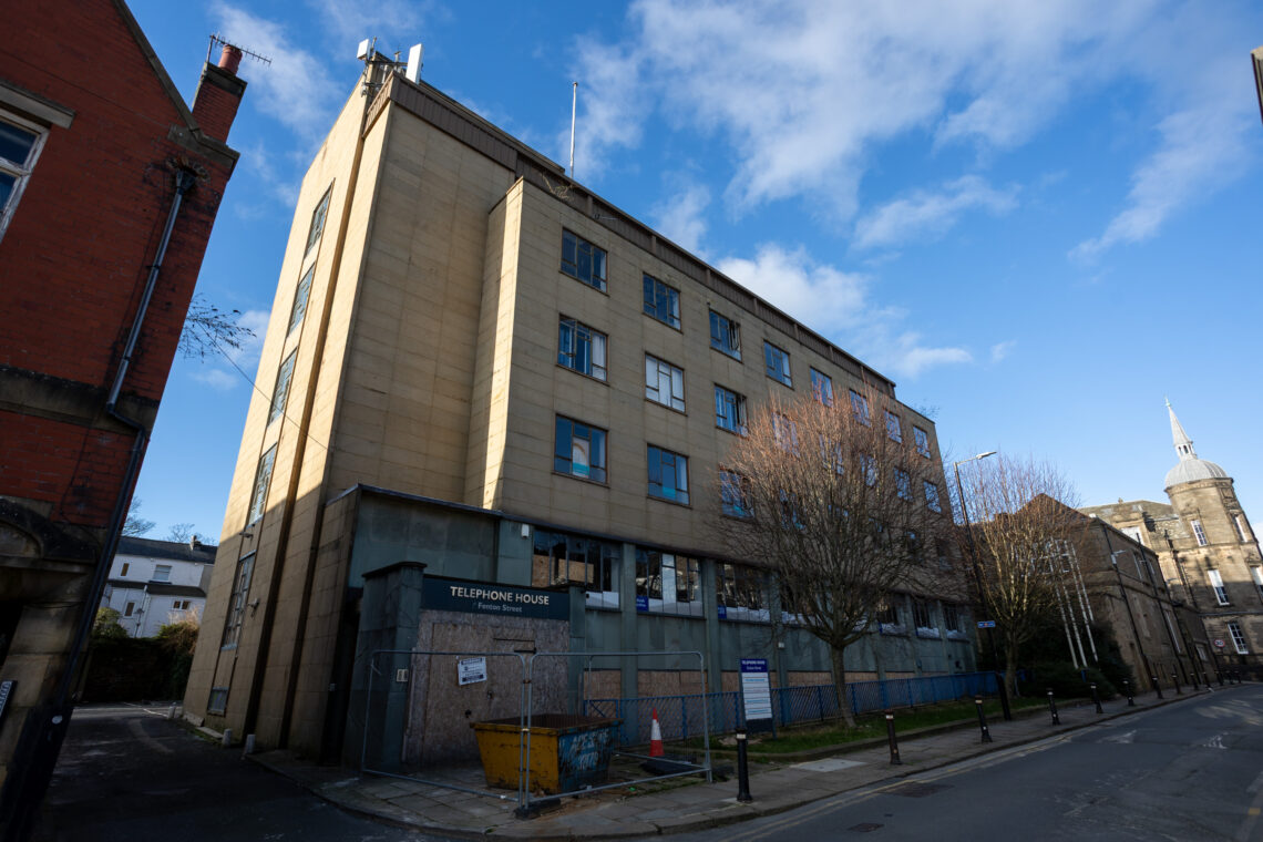 A boarded-up, beige four-storey office building labelled TELEPHONE HOUSE stands on a quiet street, with a yellow skip in front. The windows are broken or missing, and leafless trees line the pavement. The sky is partly cloudy and blue.