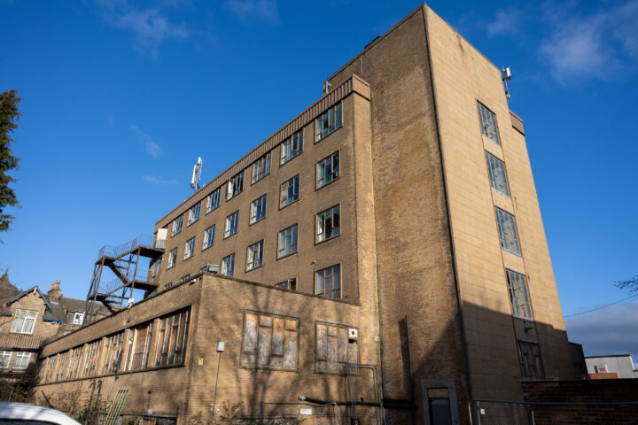 A five-storey, rectangular, tan-brick building with many windows, some boarded up, under a blue sky. A metal fire escape is attached. The building looks abandoned, and some lower windows are damaged. Neighbouring buildings and trees are partially visible.