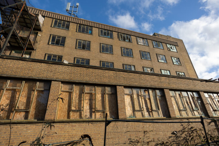 A large, abandoned brick building with boarded-up lower windows, many broken upper windows, and overgrown vegetation in front. A metal fire escape is attached to the left side. The sky is blue with clouds.