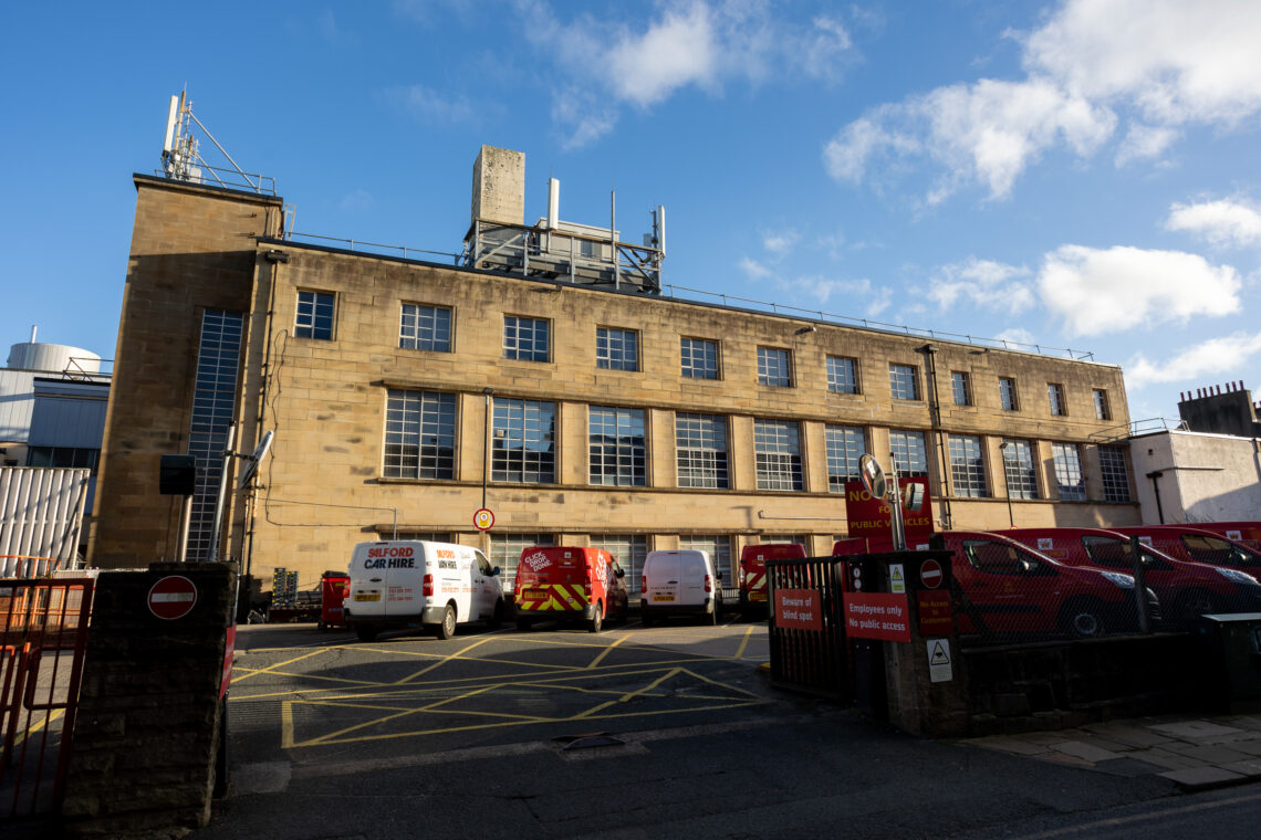 A tan brick industrial building with large rectangular windows is shown. Several red and white vans are parked in front, with yellow and white parking lines on the ground. The sky is blue with scattered clouds. Signs and barriers are also visible near the entrance.