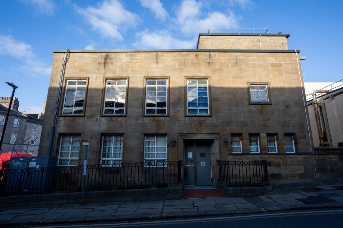 A two-storey beige stone building with nine front windows and a grey double door, metal railings, and a pavement in front. Shadows partially cover the facade. A blue sky with some clouds is visible above the building.