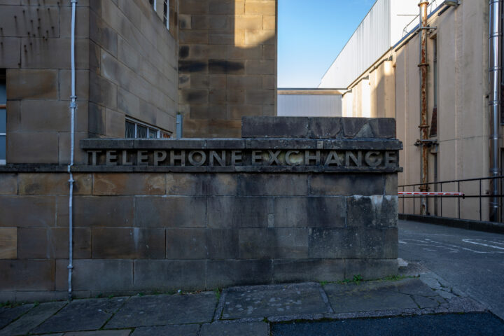 A stone wall outside a building has the words TELEPHONE EXCHANGE engraved on it. The wall is weathered, and buildings with tan and beige façades surround the area. There's a narrow passage and a barrier gate to the right.