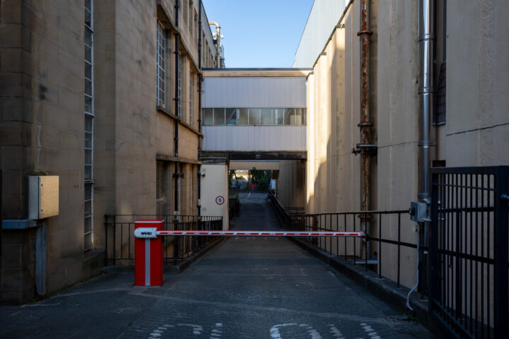 A narrow alleyway between two beige buildings with large windows, a red and white security barrier blocks entry. Overhead, a white enclosed walkway connects the buildings. The scene is lit by daylight and appears industrial.