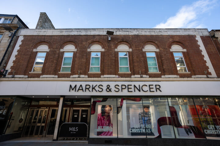 A Marks & Spencer shopfront with a brown brick façade, arched upper windows, and large display windows below featuring Christmas-themed posters. The white sign above the entrance displays the shop name in bold black letters. Blue sky is visible above.