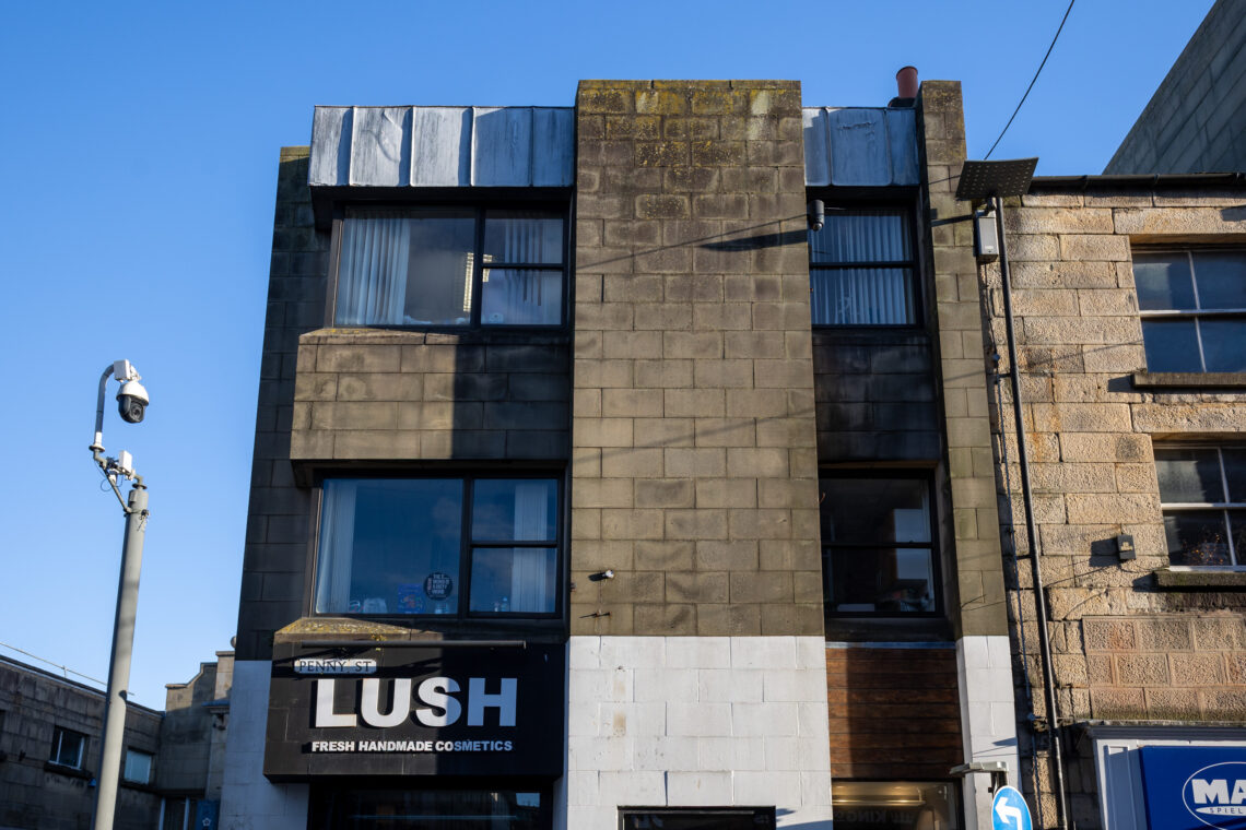 A three-storey stone building with a LUSH Fresh Handmade Cosmetics shop on the ground floor. The upper floors have large, square windows and stained, weathered walls. A CCTV camera is mounted on the left. Bright blue sky overhead.