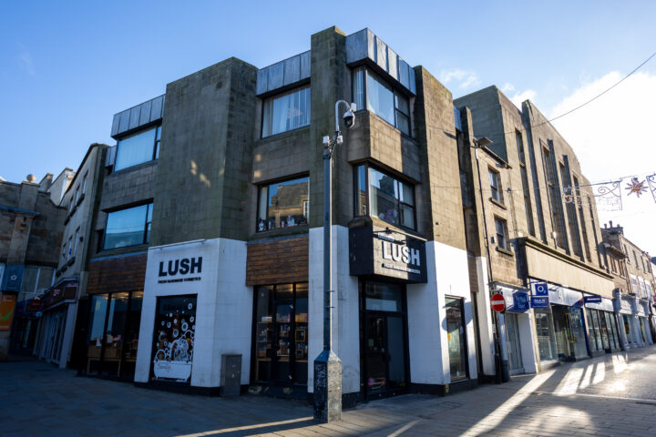Corner building with modern architecture, featuring a LUSH cosmetics shop on the ground floor. Large windows display products inside. Other shops and a bank line the street. Blue sky, sunlight casting shadows, and festive lights are visible overhead.