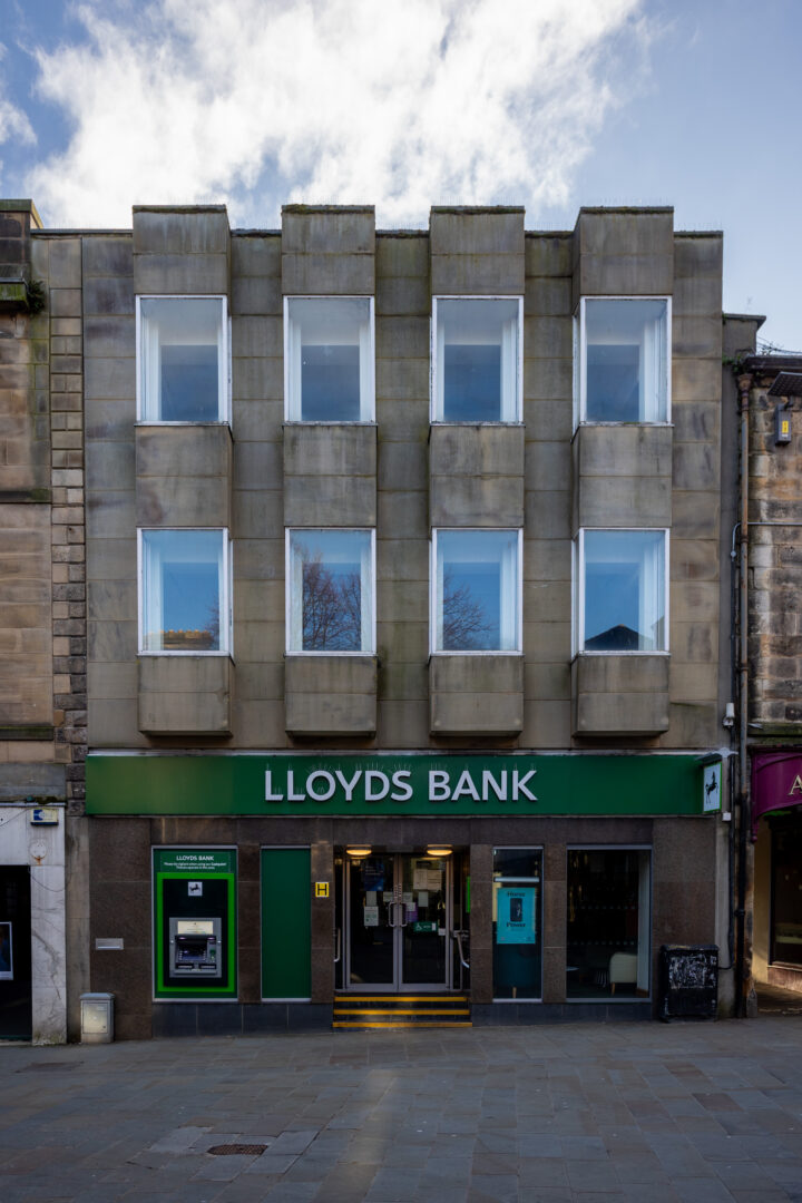 A Lloyds Bank branch with a modern stone façade and six white-framed windows above a green sign. Two cashpoints and a glass entrance door are at street level. The pavement is empty, and the sky is partly cloudy.