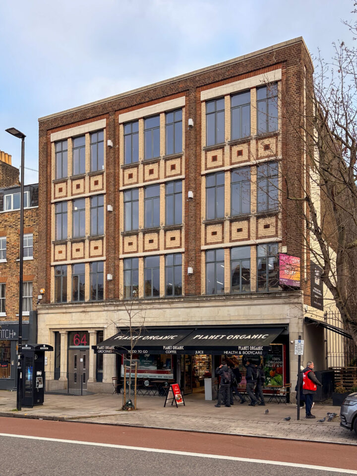 Three-storey brick building with large vertical windows, geometric brick pattern around windows, a grocery shop named Mint Organic at street level. Several people stand and walk near the entrance. Bicycles are parked outside. The number 64 is visible.