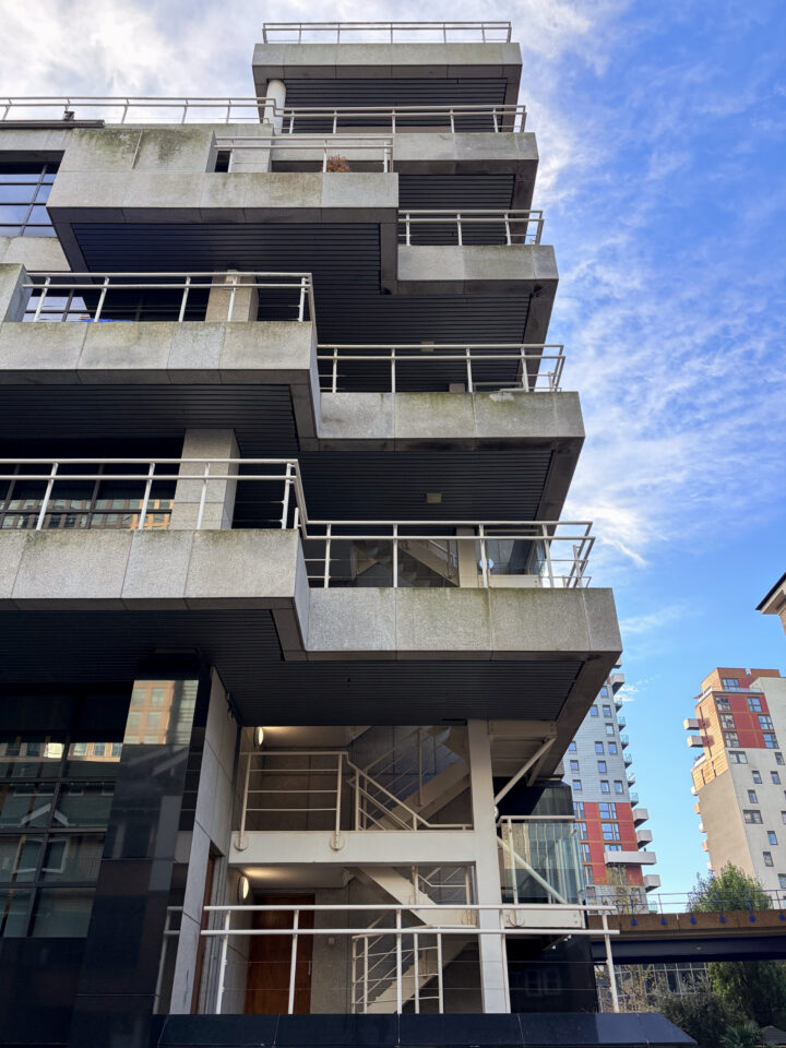 Modern concrete building with multiple staggered balconies and white railings, viewed from below. The structure has sharp angles and visible weathering. In the background, taller buildings and a blue sky with scattered clouds are visible.