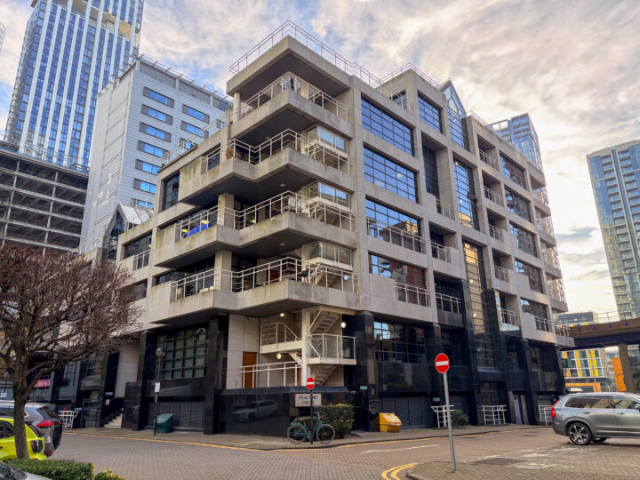 Modern multi-storey office building with large glass windows, angular balconies, and external staircases. Surrounded by high-rise buildings, parked cars, and leafless trees. Two no entry signs and a bicycle are visible near the entrance on a cloudy day.